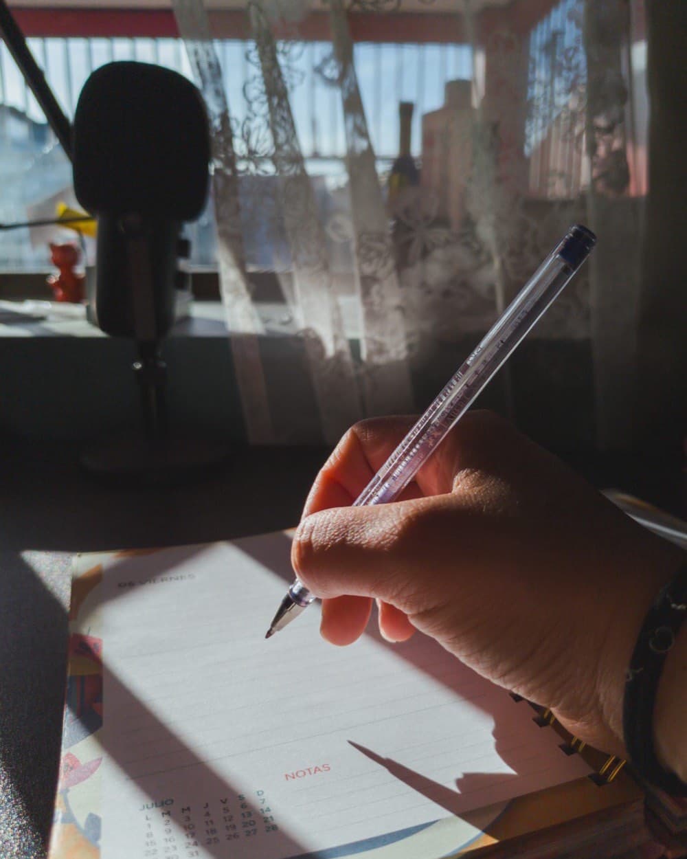 Une main écrit au stylo sur un carnet ligné, un microphone posé en arrière-plan